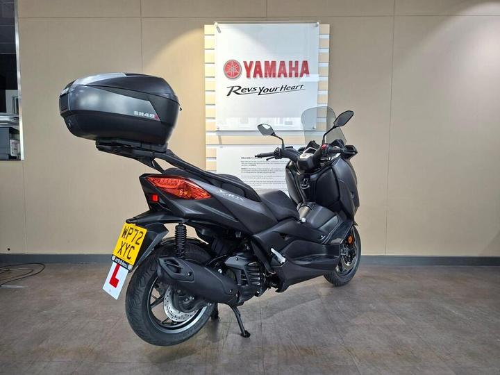 A black Yamaha scooter with a top case is displayed in a showroom, featuring a "Yamaha Rev's Your Heart" sign in the background. The scooter has a learner driver plate attached and is positioned on a smooth floor.