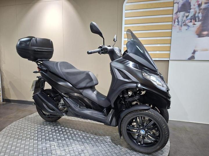 A black three-wheeled scooter with a sleek design, featuring a comfortable seat and a rear storage box, displayed on a round platform in a modern indoor setting.