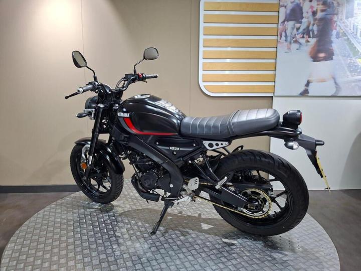 Yamaha motorcycle displayed on a circular metallic platform, with a black and red color scheme, featuring a sleek design and retro style, against a beige wall background.