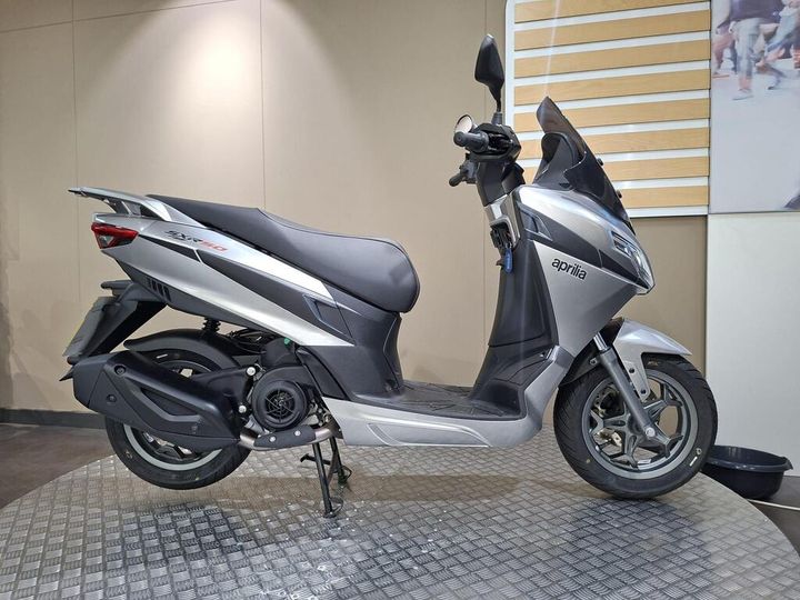 Aprilia SXR 50 2026 A silver Aprilia scooter displayed on a textured surface against a neutral background.