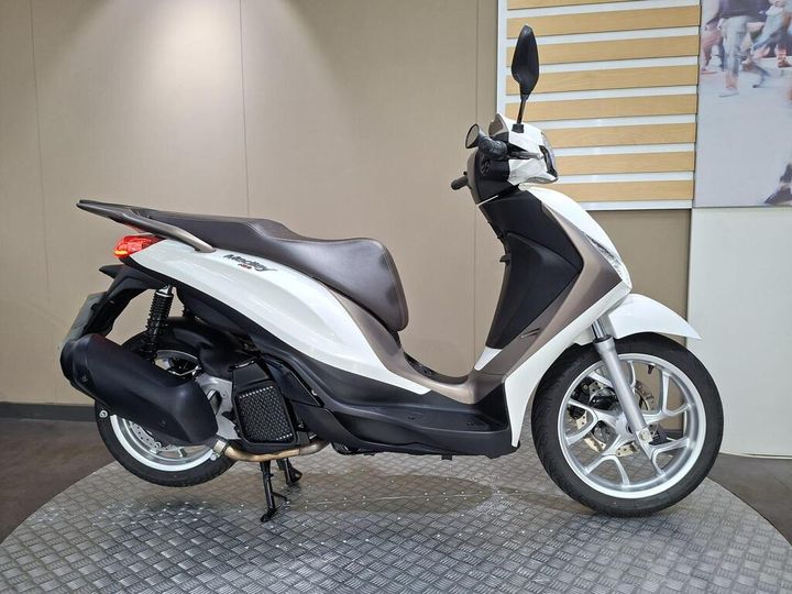 Piaggio Medley (2025) A white scooter with a brown seat, displayed on a circular platform, against a neutral background. The scooter features a modern design, with a prominent front headlight and sleek body contours.
