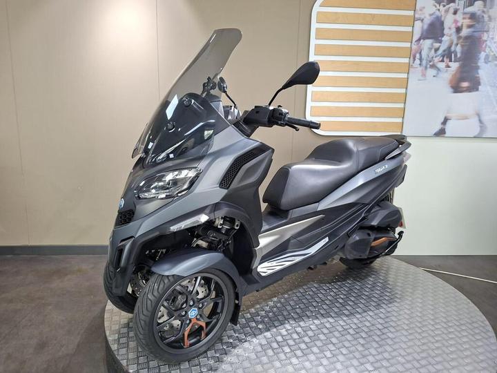 A black and silver three-wheeled scooter displayed on a round metallic platform, featuring a large windshield and modern design elements. The background is a subtle light tan color with a partially visible textural wall.