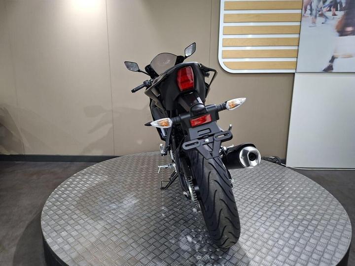 A rear view of a black motorcycle on a circular metallic platform, showcasing its tail lights, exhaust, and tire tread pattern against a neutral background.