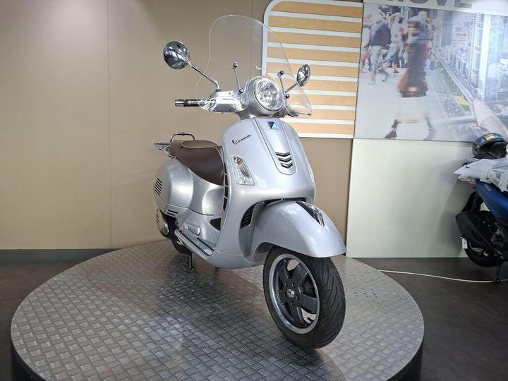 A silver Vespa scooter displayed on a circular stand in an indoor showroom, with a clear windshield and a brown seat, against a neutral background.