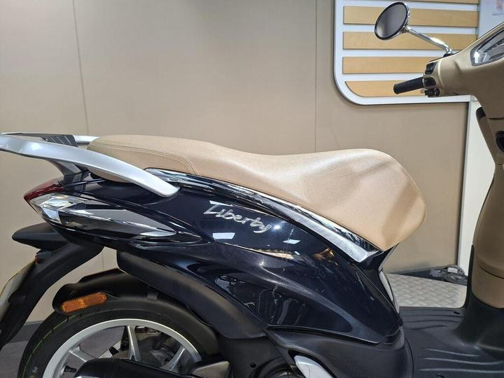 A close-up view of a blue scooter with a light beige seat, featuring the word "Liberty" in white lettering on the side.