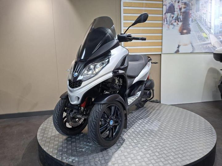 A three-wheeled scooter with a sleek silver and black design, displayed on a circular metal platform in a modern setting. The scooter features a prominent windshield and is positioned against a neutral background with a blurred image of people and buildings.