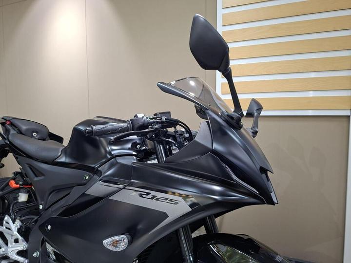 A close-up view of the front section of a black motorcycle, highlighting the handlebars, windshield, and side mirrors against a minimalist background.