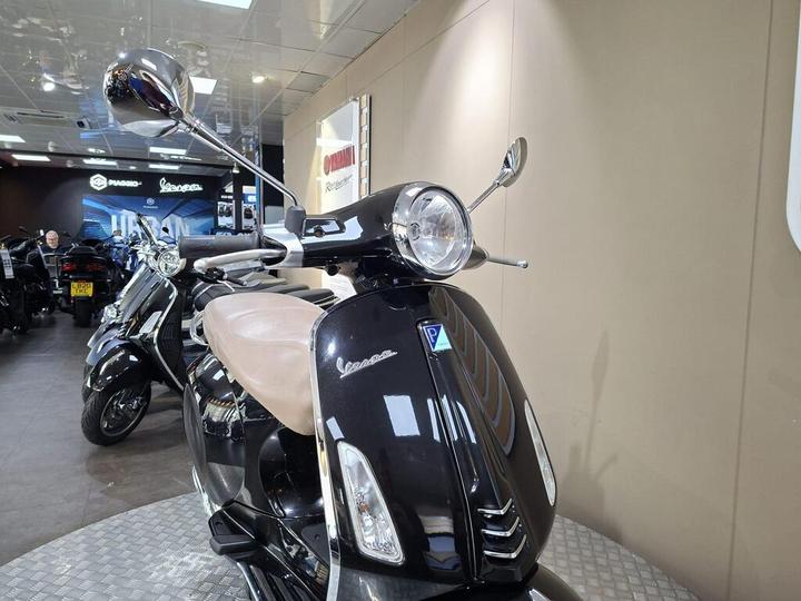 A black Vespa scooter with a tan seat displayed in a modern showroom, featuring chrome mirrors and a shiny headlight, with other scooters in the background.