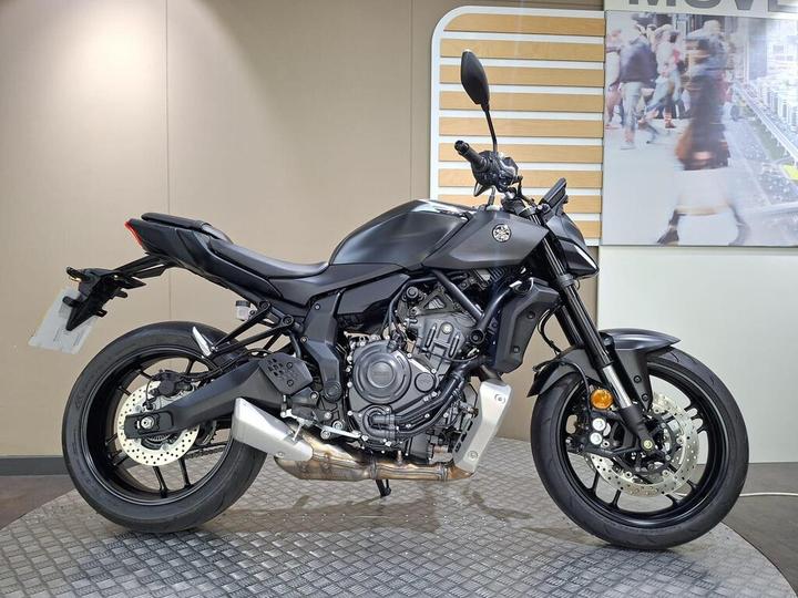 Yamaha MT-07 2026 A black motorcycle is displayed on a textured surface with a minimalistic background featuring a panel and a graphic of people walking. The bike's sleek design includes a sporty frame, prominent exhaust, and modern styling elements.