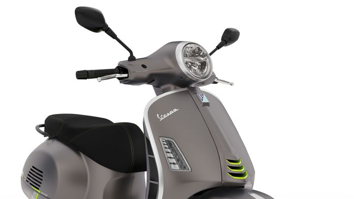 A close-up view of a modern grey Vespa scooter featuring a round headlight, handlebar controls, and a stylish seat with green stitching.