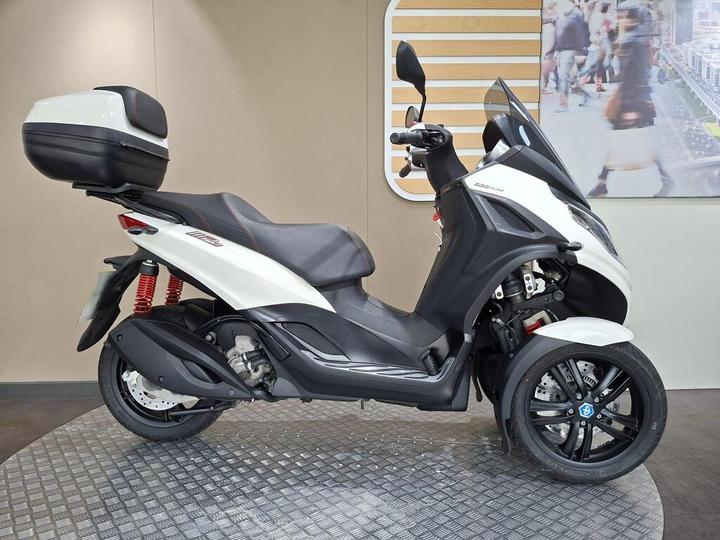 Piaggio MP3 300 Sport (2021) A white three-wheeled scooter with a black seat and a top case, positioned on a textured surface against a neutral background with abstract designs.