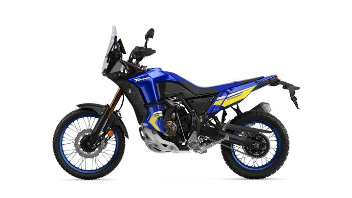 A blue and black adventure motorcycle with yellow accents, featuring off-road tires, a tall windshield, and a rugged design, positioned against a white background.