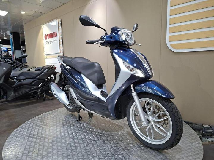 A blue and silver scooter is displayed on a diamond-patterned platform in a showroom, with a Yamaha sign visible in the background and another motorcycle partially shown to the left.