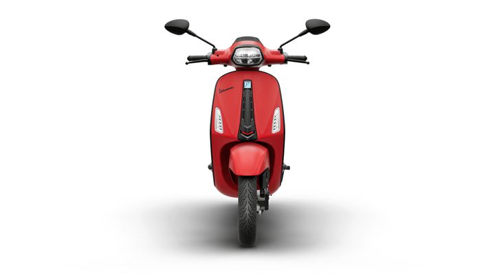A red scooter with a sleek design, facing forward, featuring a prominent headlight, mirrors, and a distinctive brand logo.