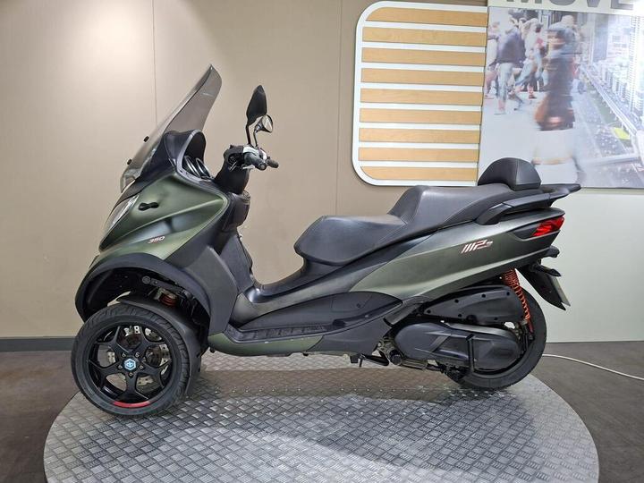 A dark green three-wheeled scooter displayed on a circular platform, featuring a sleek design, black wheels, and a transparent windshield, set against a neutral background with a modern wall graphic.