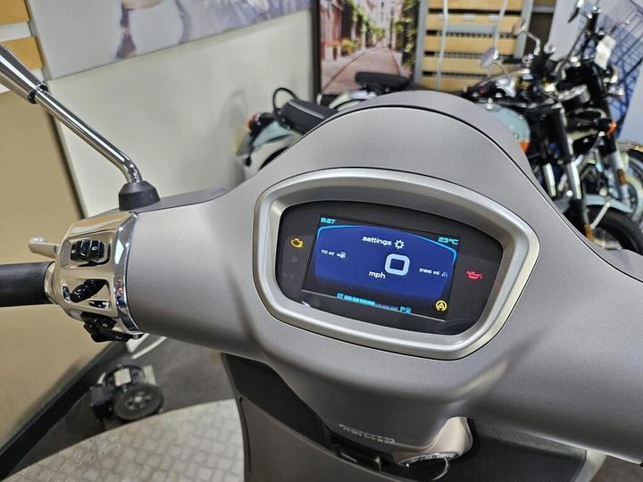 A close-up view of a scooter dashboard featuring a digital display showing settings, speedometer indicating 0 mph, and a temperature reading of 23°C, with chrome accents and handlebar controls visible in the foreground.