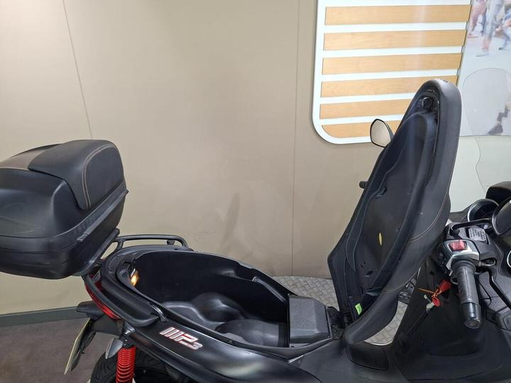 A close-up view of a scooter with its seat lifted, revealing storage space underneath. The scooter features a black design with a label "MP3" visible on the side. The background is neutral with minimal decor.