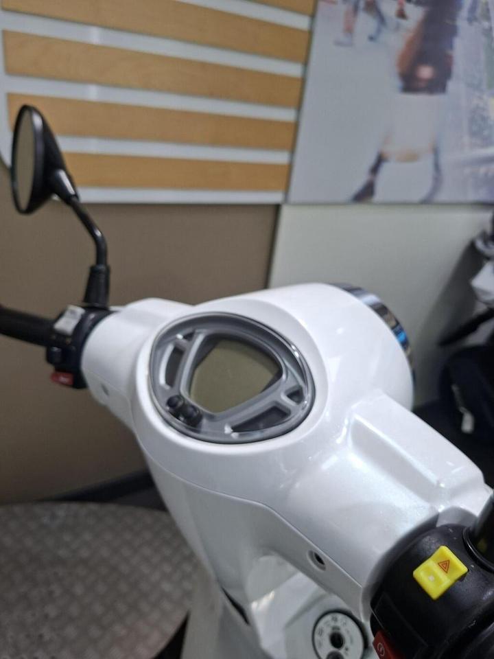 Close-up of a white scooter's handlebar and dashboard, featuring a digital display and control buttons, with a blurred background.