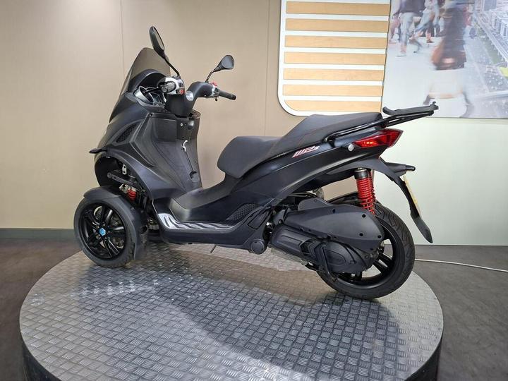 A black three-wheeled scooter displayed on a circular platform, showcasing a sleek design with a comfortable seat, front windshield, and a modern dashboard. The background features a subtle design, enhancing the focus on the scooter.