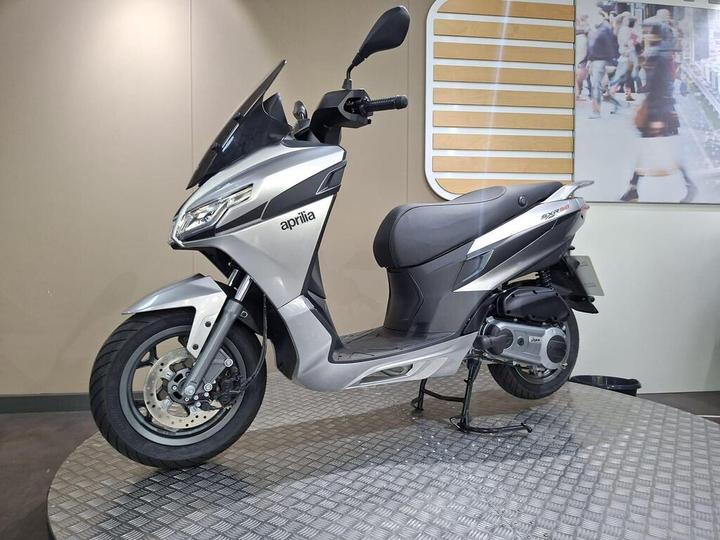 A silver Aprilia scooter showcased on a textured display platform, set against a light-colored wall with a blurred background image of people walking.