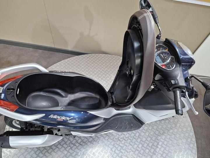 A top-down view of a scooter, showcasing its open seat compartment and dashboard area, with a grey checkered floor beneath.