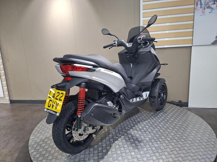 A modern three-wheeled scooter in silver and black, featuring red suspension springs, positioned on a textured surface with a plain background.