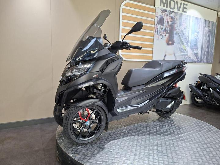 A black three-wheeled scooter displayed in a showroom, featuring a large windscreen and sporty design, with a checkered floor and a blurred background of more scooters and a promotional wall.