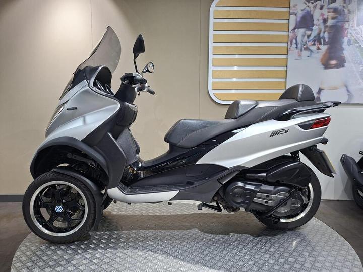 A side view of a silver three-wheeled scooter with a black seat, displayed on a metal platform against a neutral background.