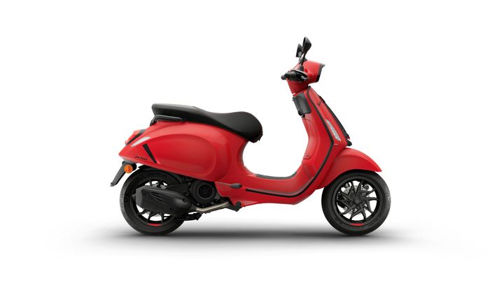 A sleek red scooter is shown from the side, featuring a smooth body design, a comfortable black seat, and stylish wheels. The background is plain white, highlighting the scooter's vibrant color.