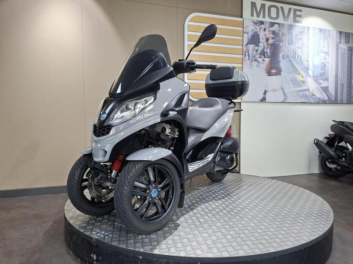 A gray three-wheeled scooter displayed on a circular platform in a showroom, with a modern background featuring images of city life.
