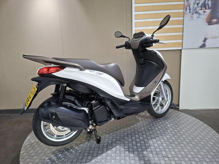 A sleek, modern scooter in white and brown is displayed at an angle, showcasing its curved design and rear view on a textured platform against a neutral background.