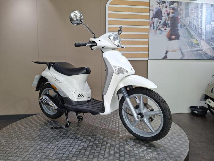A white scooter displayed on a round platform, featuring a prominent front headlight, rearview mirrors, and a comfortable seat, against a neutral background with pedestrian imagery.