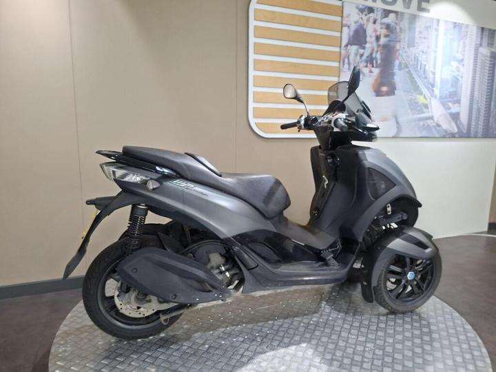 A sleek, gray three-wheeled scooter displayed in a showroom setting, featuring modern design elements and a minimalistic background.