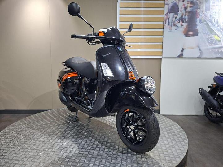 A black Vespa scooter with orange accents is displayed on a circular, textured platform in a showroom. The background features a partially visible wall and a blurred image of pedestrians.