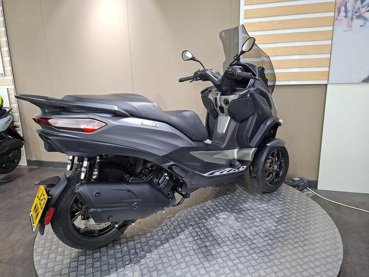 A sleek, gray three-wheeled scooter positioned on a circular display platform in a showroom, showcasing its modern design and features.