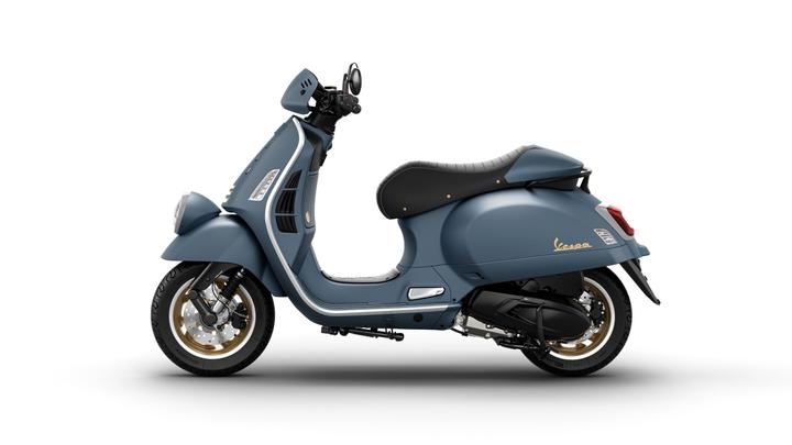 A modern blue Vespa scooter with a sleek design, featuring a black seat, gold accents on the wheels, and a classic handlebar setup, shown from a side view.