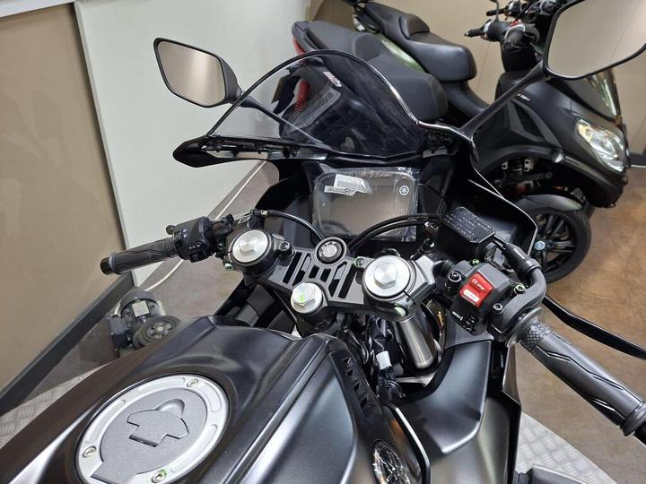 Close-up view of a motorcycle's handlebars, featuring a digital display, control buttons, and mirrors, with additional parked scooters visible in the background.