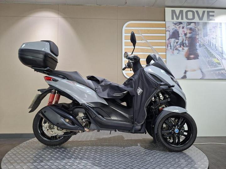 Piaggio MP3 300 Sport 2021 A grey three-wheeled scooter with a side storage compartment, displayed on a circular platform in a modern showroom setting, featuring a beige wall with a "MOVE" sign in the background.
