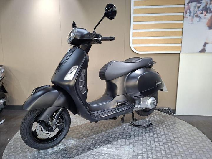 A sleek black scooter is showcased from the side, featuring a round front headlight and a comfortable seat, set against a neutral background with a textured display surface.