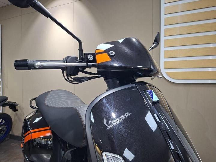 Close-up view of a Vespa scooter's handlebar and display, showcasing a sleek black design with orange accents and modern controls, set against a neutral background.