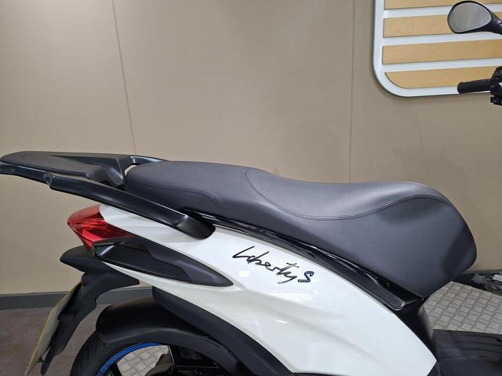 A side view of a black and white scooter, featuring a sleek design with the name "Liberty S" written on the body. The seat is modern and contoured, positioned above the rear wheel and set against a neutral background.