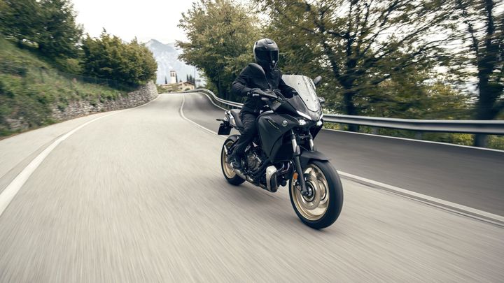 A motorcyclist in black gear rides a sleek motorcycle on a winding road, surrounded by trees and hills, with a sense of motion captured in the image.