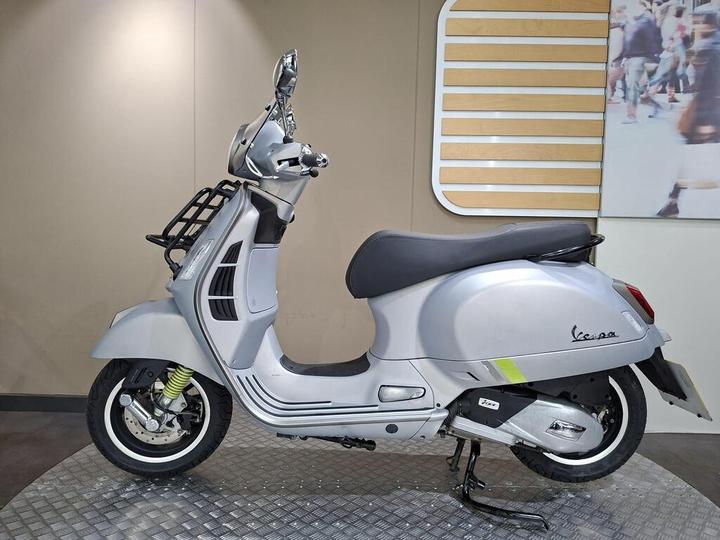 A silver Vespa scooter displayed on a textured platform, with a minimalist background featuring wooden panels. The scooter features a black seat, a front basket, and a prominent logo on the rear.