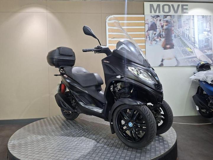 A three-wheeled black scooter displayed on a circular platform in a showroom, featuring a windshield, a comfortable seat, and a rear storage box. The backdrop includes a "MOVE" sign and a blurred cityscape image.