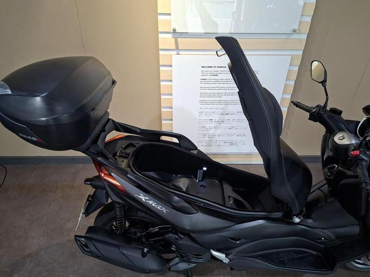 A black Yamaha Xmax scooter is displayed with its seat lifted, revealing a storage compartment. A black top case is mounted on the rear, and a wall behind the scooter features informational text.