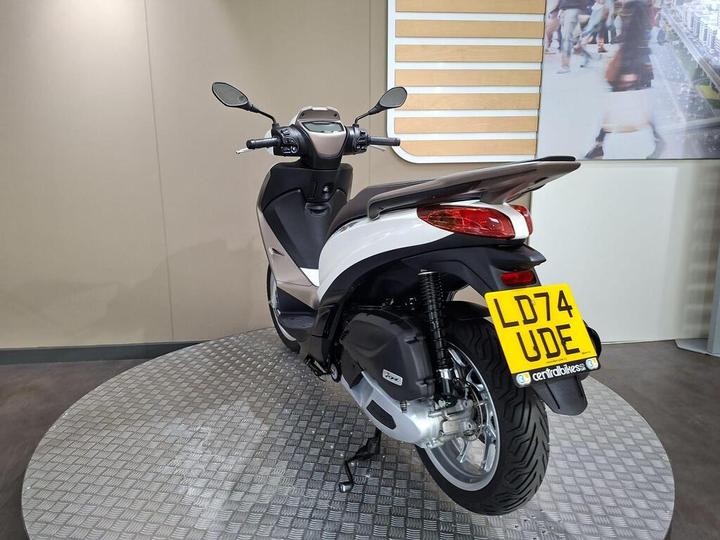 A rear view of a modern scooter displayed on a circular platform, featuring a gray and white color scheme, with its license plate visible reading "LD74 UDE." The background consists of a neutral wall and a digital image of people walking.