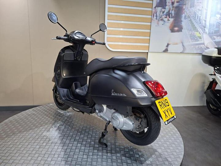 A matte black Vespa scooter positioned on a circular metal platform, with its right side facing the viewer. The background features a beige wall and an abstract image of a cityscape, while the scooter showcases its distinctive shape, mirrors, and license plate.