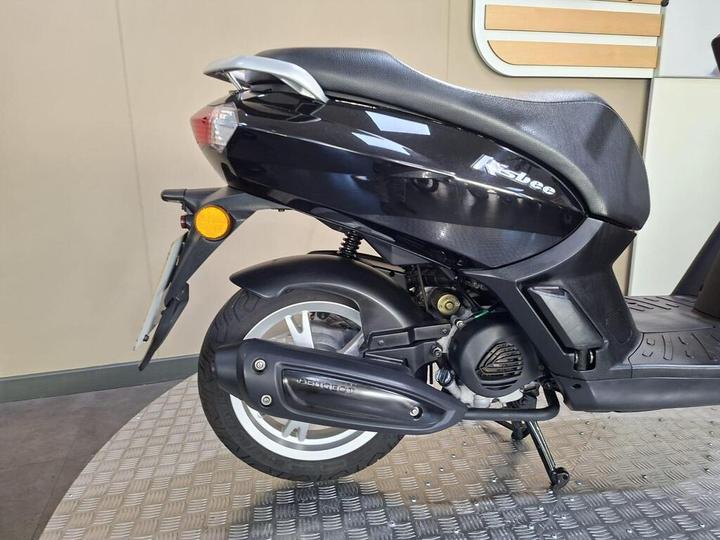 A close-up view of the rear of a black scooter, featuring a rounded tail light, a sleek body design, and a visible exhaust system. The background includes a light-colored wall and a textured floor.