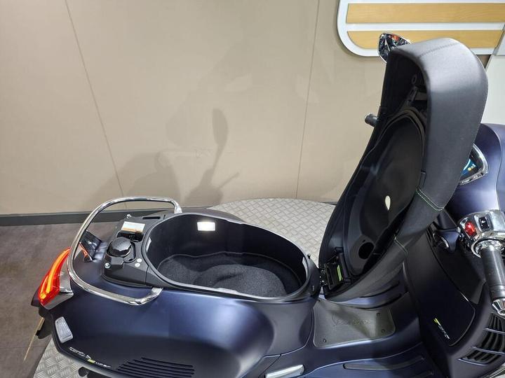 A close-up view of a scooter's storage compartment with the seat slightly lifted, showing a black, padded interior and controls on the side. The background features a neutral-colored wall.