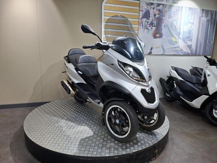 A silver three-wheeled scooter displayed on a circular platform in a showroom, with a modern design and a clear windshield. In the background, there is a wall featuring a cityscape image, and another scooter is visible to the right.